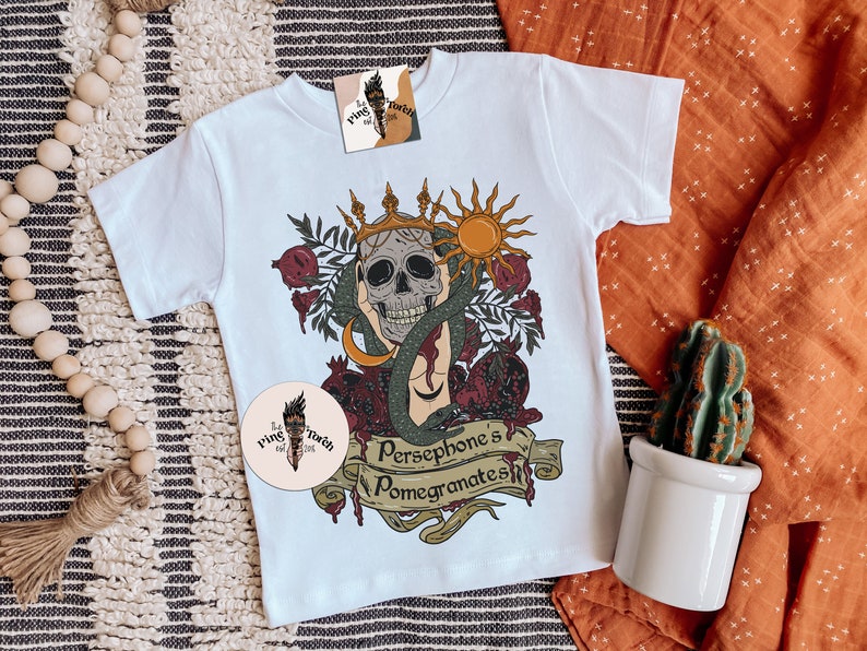 May include: White t-shirt featuring a graphic of a skull wearing a crown, surrounded by pomegranates and a snake. The text "Persephone's Pomegranates" is written on a banner below the skull.