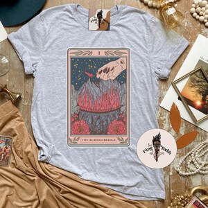 May include: Gray t-shirt with a tarot card design featuring a hand reaching towards a burning bridge. The card is titled "The Burned Bridge".