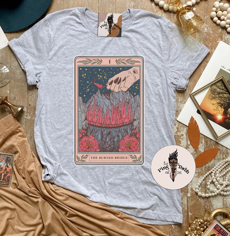 May include: Gray t-shirt with a tarot card design featuring a hand reaching towards a burning bridge. The card is titled "The Burned Bridge".