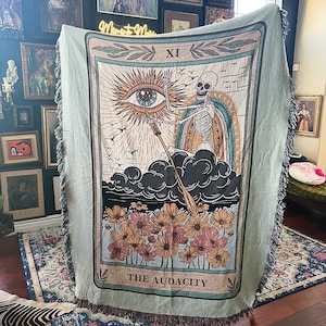 May include: A woven tapestry featuring a tarot card design. The card is the eleventh card, "The Audacity." The card depicts a skeleton holding a staff with an eye at the top. The background is a blue sky with clouds and a sun with an eye. The card is surrounded by a green border with a floral design.