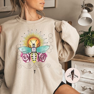 May include: A beige sweatshirt with a graphic print of a bee with a sun and honeycomb design. The bee is surrounded by pink roses and the text "Queen" is on the sun.
