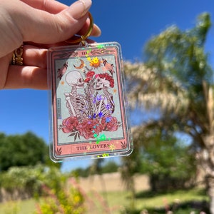 May include: A clear keychain with a pink and white tarot card design. The card features a skeleton couple holding hands with flowers and stars. The card reads "III THE LOVERS".