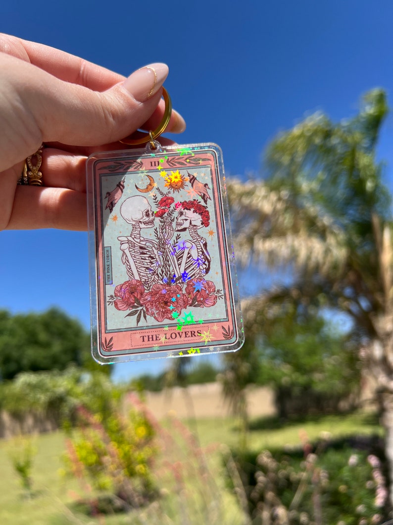 May include: A clear keychain with a pink and white tarot card design. The card features a skeleton couple holding hands with flowers and stars. The card reads "III THE LOVERS".