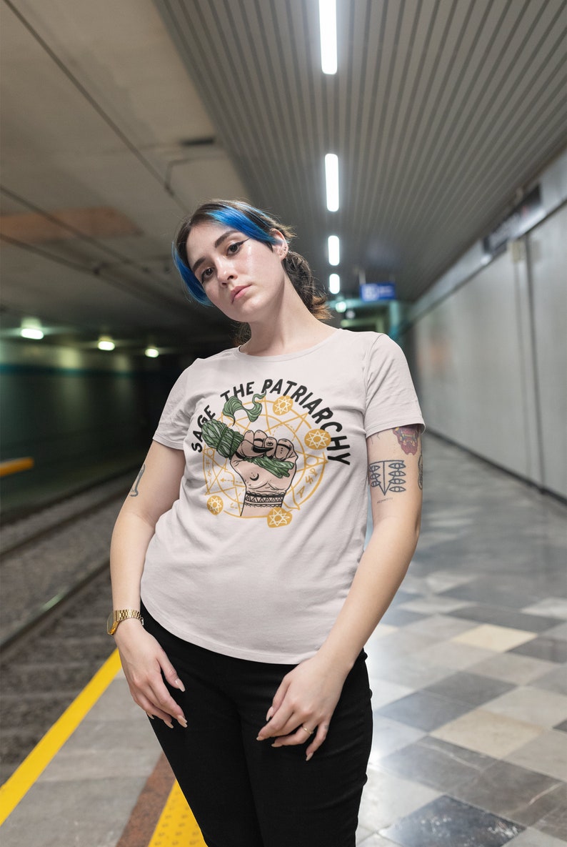 Puede incluir: Una mujer con una camiseta blanca con un gr&aacute;fico de una mano sosteniendo un manojo de salvia y el texto "Sage the Patriarchy".
