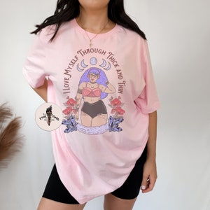 Puede incluir: Camiseta rosa con un gr&aacute;fico de una mujer con cabello morado y un sujetador negro, rodeada de lunas y cristales. El texto "I love myself through thick and thin" est&aacute; impreso alrededor del gr&aacute;fico.