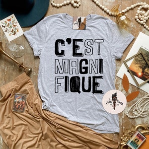 May include: A gray t-shirt with the French phrase "C'est Magnifique" printed in black. The shirt is laid out on a wooden surface with various decorative items around it.