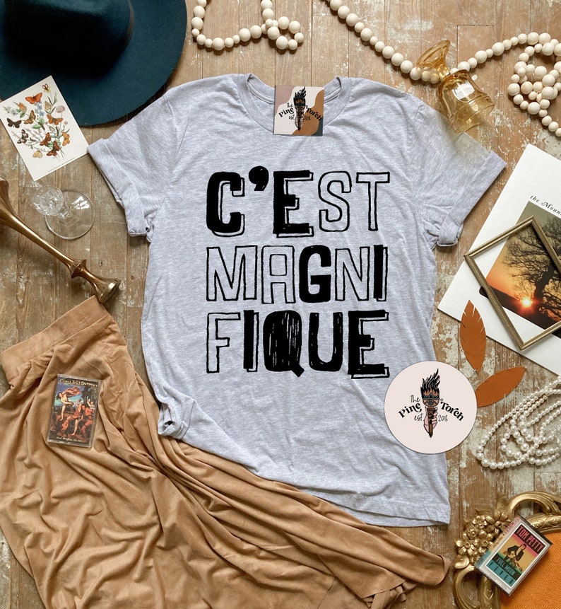 May include: A gray t-shirt with the French phrase "C'est Magnifique" printed in black. The shirt is laid out on a wooden surface with various decorative items around it.