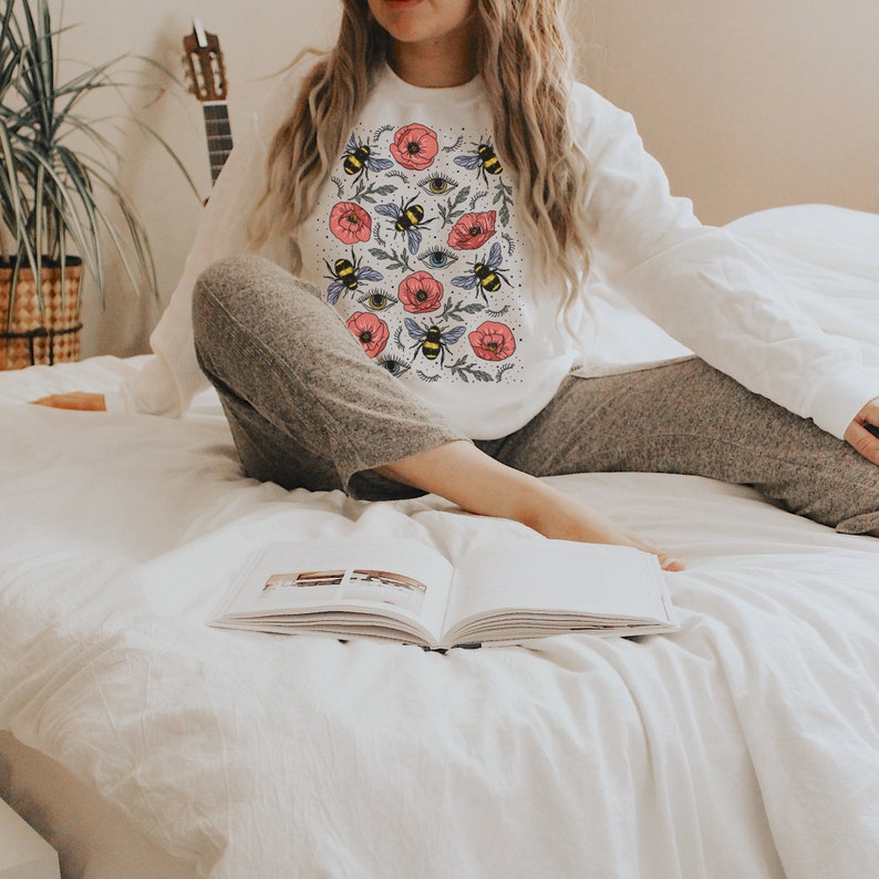 K&ouml;nnte beinhalten: Ein wei&szlig;er Pullover mit einem bunten Druck von Bienen, Blumen und Augen. Der Pullover wird von einer Person getragen, die auf einem wei&szlig;en Bett mit einem Buch sitzt.