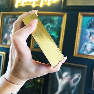 May include: A close-up of a deck of playing cards with a gold foil finish. The cards are held in a hand, with the edges of the cards visible.