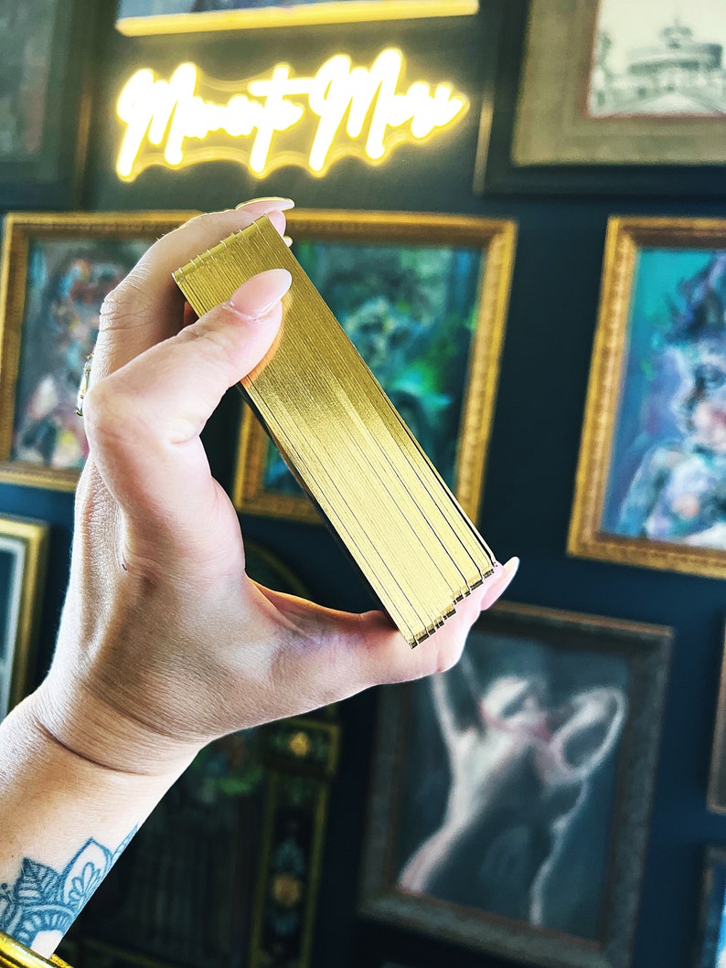May include: A close-up of a deck of playing cards with a gold foil finish. The cards are held in a hand, with the edges of the cards visible.