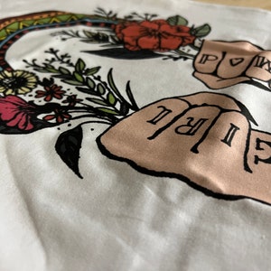 May include: White t-shirt with a colorful rainbow design and two fists with the words "GIRL" and "POWER" written on the knuckles. The text "THE PINE TORCH" is printed on the bottom right corner of the shirt.