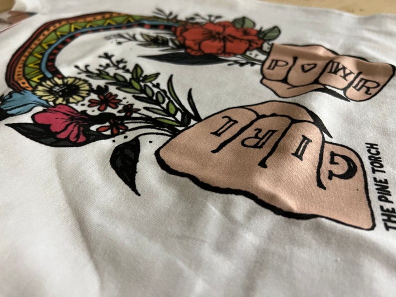 May include: White t-shirt with a colorful rainbow design and two fists with the words "GIRL" and "POWER" written on the knuckles. The text "THE PINE TORCH" is printed on the bottom right corner of the shirt.