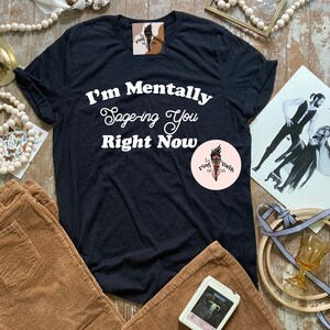 May include: Black t-shirt with white text that reads "I'm Mentally Sage-ing You Right Now". The shirt is on a table with brown corduroy pants, a white cassette tape, and a white and brown beaded garland.
