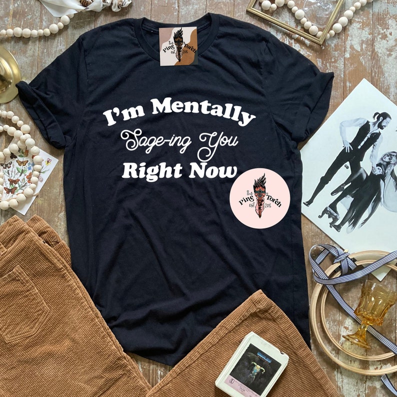 May include: Black t-shirt with white text that reads "I'm Mentally Sage-ing You Right Now". The shirt is on a table with brown corduroy pants, a white cassette tape, and a white and brown beaded garland.