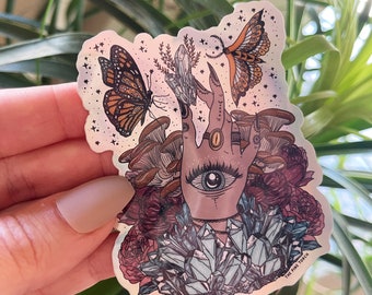May include: A holographic sticker featuring a hand with an all-seeing eye, surrounded by crystals, mushrooms, and butterflies. The sticker has a pink, purple, and blue color scheme and includes the text "The Pink Torch".