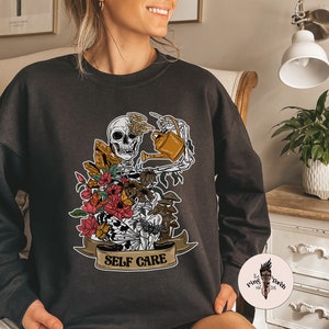 May include: Black sweatshirt with a graphic of a skeleton watering plants with the text "Self Care" on a banner.