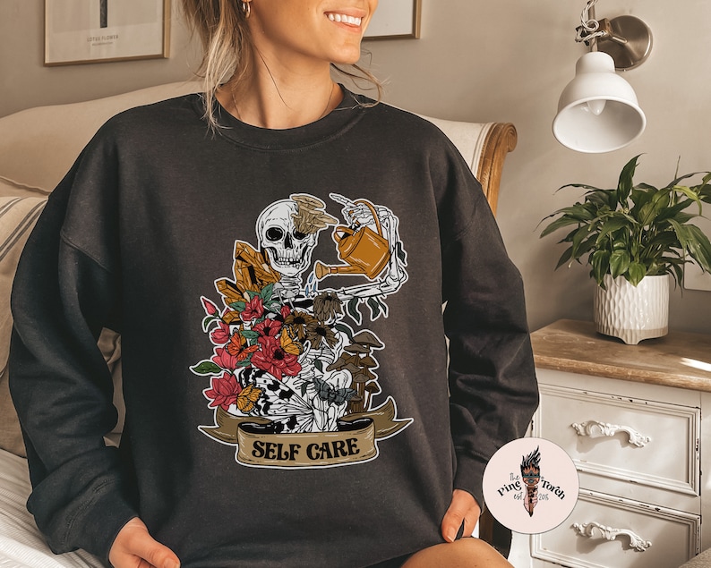 May include: Black sweatshirt with a graphic of a skeleton watering plants with the text "Self Care" on a banner.