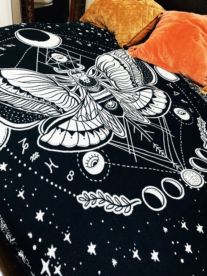 May include: Black and white tapestry featuring a moth with intricate details, surrounded by moons, stars, and eyes. The tapestry has a geometric design and a celestial theme.