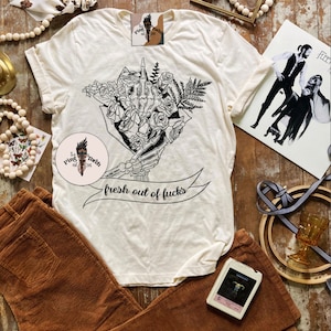 K&ouml;nnte beinhalten: Cremefarbenes T-Shirt mit einer schwarzen Grafik einer Skeletthand, die Blumen h&auml;lt, mit dem Text "fresh out of fucks" auf einem Banner. Braune Cordhose ist darunter gefaltet. Ein Vintage-Album und eine Kassette sind ebenfalls sichtbar.