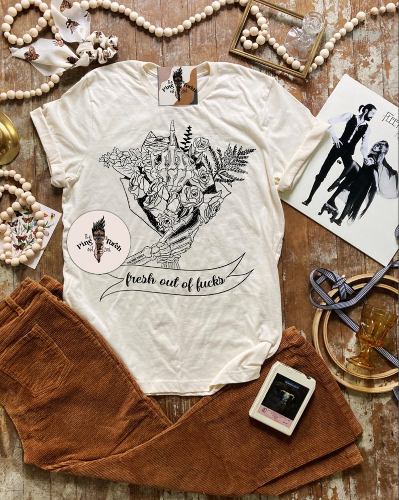 K&ouml;nnte beinhalten: Cremefarbenes T-Shirt mit einer schwarzen Grafik einer Skeletthand, die Blumen h&auml;lt, mit dem Text "fresh out of fucks" auf einem Banner. Braune Cordhose ist darunter gefaltet. Ein Vintage-Album und eine Kassette sind ebenfalls sichtbar.
