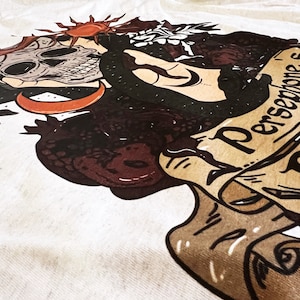 May include: A white t-shirt featuring a black and brown illustration of a skull, a snake, and a crescent moon. The text "Persephone's" is written on a scroll.