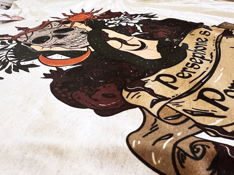 May include: A white t-shirt featuring a black and brown illustration of a skull, a snake, and a crescent moon. The text "Persephone's" is written on a scroll.