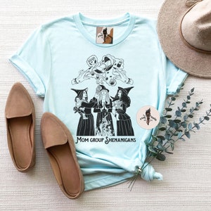 K&ouml;nnte beinhalten: Hellblaues T-Shirt mit einer schwarz-wei&szlig;en Illustration von drei Hexen mit einem Kessel und dem Text "Mom Group Shenanigans".