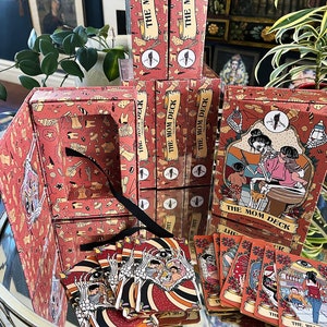 May include: A set of playing cards with a red and white design featuring a skeleton family. The cards are in a box with a red and white design featuring a skeleton family. The box is open and the cards are spilling out. The text on the box reads "The Mom Deck".