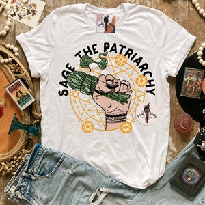 Puede incluir: Camiseta blanca con un dise&ntilde;o gr&aacute;fico verde y amarillo que dice "Sage the Patriarchy". El gr&aacute;fico presenta una mano sosteniendo un manojo de salvia y un c&iacute;rculo con estrellas y soles.