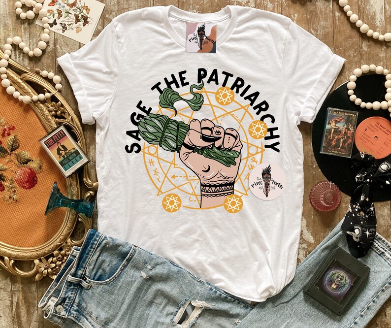 Puede incluir: Camiseta blanca con un dise&ntilde;o gr&aacute;fico verde y amarillo que dice "Sage the Patriarchy". El gr&aacute;fico presenta una mano sosteniendo un manojo de salvia y un c&iacute;rculo con estrellas y soles.