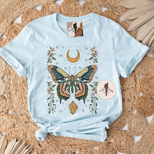 May include: Light blue t-shirt with a colorful butterfly graphic. The butterfly has large eyes and is surrounded by flowers, stars, and a crescent moon. The text "The Pine & Feather Co." is on a small circle in the bottom right corner.