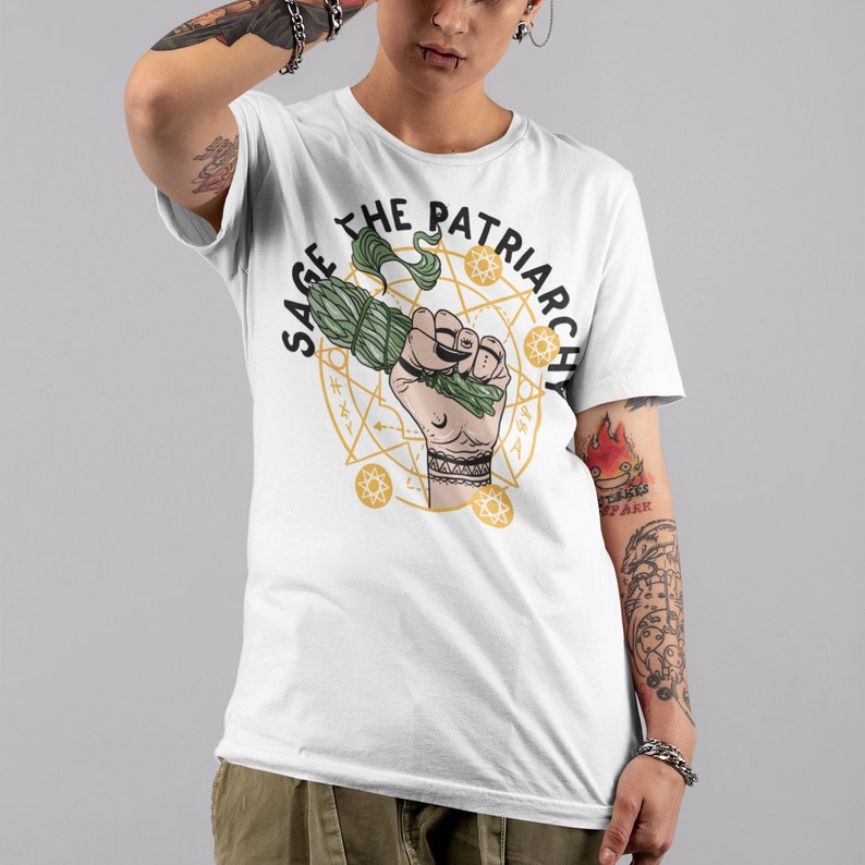 Puede incluir: Camiseta blanca con un gr&aacute;fico de un pu&ntilde;o sosteniendo un manojo de salvia, rodeado de un c&iacute;rculo dorado con el texto "Sage the Patriarchy".