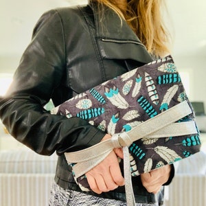 Könnte beinhalten: Eine graue Clutch mit einem silbernen Band und einem Muster aus blaugrünen und weißen Federn. Die Clutch wird von einer Person in einer schwarzen Lederjacke gehalten.
