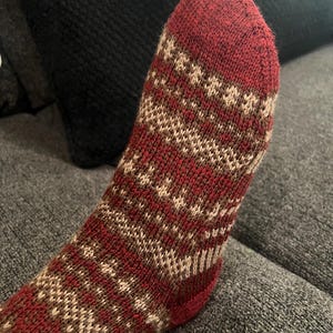 May include: A close-up of a knitted sock in shades of burgundy, brown, and cream. The sock features a geometric pattern and is made of a soft, textured material. The toe and heel are a solid burgundy color.