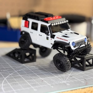 Op de afbeelding: Een witte speelgoed Jeep Wrangler Rubicon met zwarte wielen en een zwart dakrek. Het model staat op zwarte platforms. Het woord "RUBICON" staat op de motorkap.