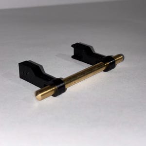 May include: A black plastic component with a gold-colored metal rod. The rod is held in place by two black brackets. The object appears to be a small mechanical part or component, possibly for a larger device.