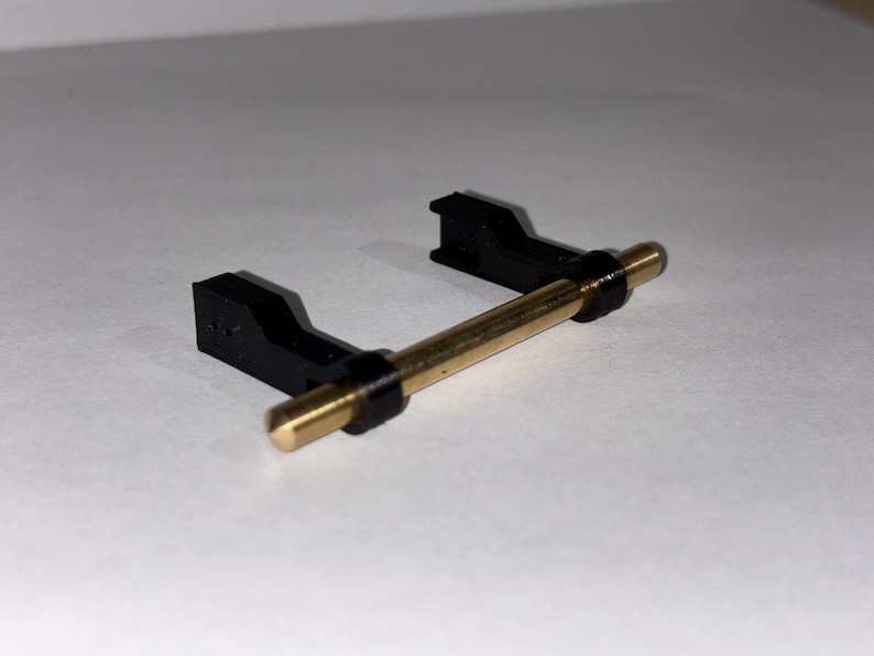 May include: A black plastic component with a gold-colored metal rod. The rod is held in place by two black brackets. The object appears to be a small mechanical part or component, possibly for a larger device.