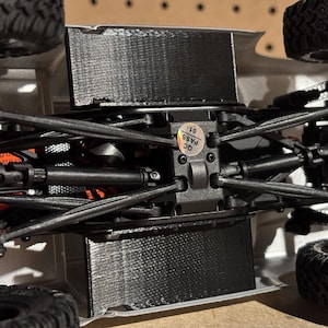 Puede incluir: Vista inferior de un chasis de camión RC negro y plateado. La imagen muestra la suspensión, los ejes y otros componentes mecánicos. Es visible una pegatina con "QC PASS 01". Los neumáticos son negros y tienen un dibujo robusto.