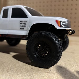 May include: A white model pickup truck with black wheels and tires. The truck has a detailed front grill and headlights. The side of the truck has sponsor logos and the text "BASE CAMP".