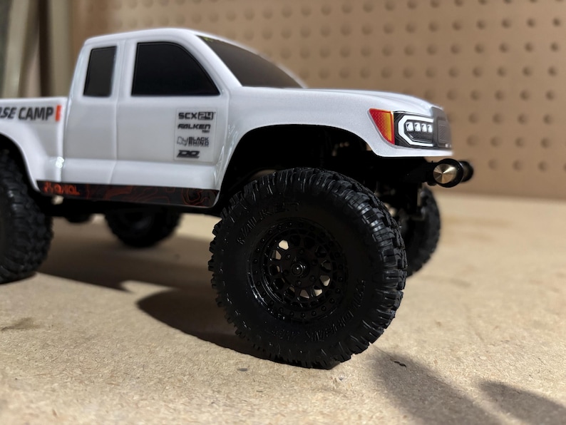 May include: A white model pickup truck with black wheels and tires. The truck has a detailed front grill and headlights. The side of the truck has sponsor logos and the text "BASE CAMP".
