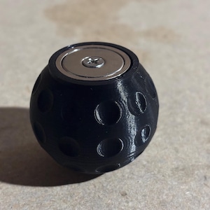 Op de afbeelding: Een zwarte bol met een getextureerd oppervlak en een zilveren metalen schijf bovenop. De bol heeft een gegraveerd patroon.