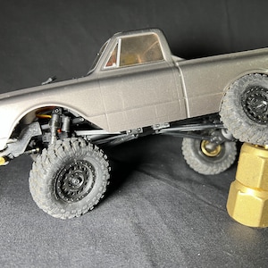Può includere: Un camioncino giocattolo grigio con ruote nere e un supporto in metallo dorato. Il camioncino è sollevato da terra e le ruote sono in aria.