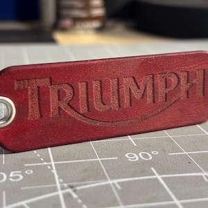May include: A red leather keychain with the word "TRIUMPH" embossed on it. The keychain has a silver-colored metal grommet for attaching to keys. The background is a gray cutting mat.