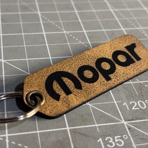 May include: A rectangular leather keychain with the word "Mopar" in black lettering. The keychain has a gold-brown textured surface and a silver key ring attached.