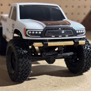 Puede incluir: Un camión de juguete blanco y negro con neumáticos negros grandes. El camión tiene un parachoques dorado y una parrilla negra. La parte delantera del camión tiene una serie de luces. El camión es una réplica en miniatura.