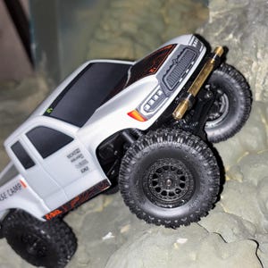 May include: A white and black toy truck with large black tires is angled on a rocky surface. The truck has orange and black accents, and the words "BASE CAMP" are visible on the side. The truck is designed to look like a real off-road vehicle.