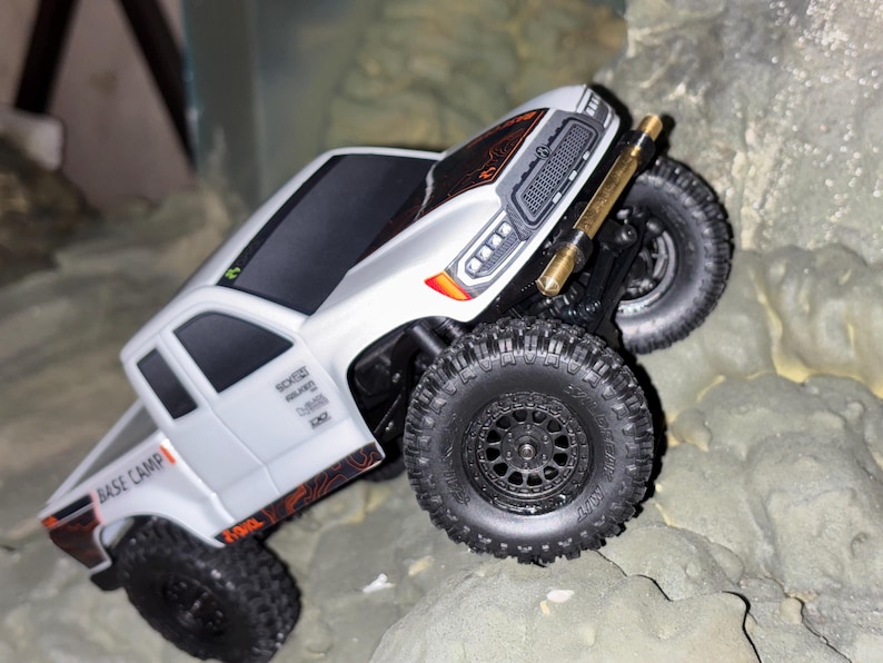 May include: A white and black toy truck with large black tires is angled on a rocky surface. The truck has orange and black accents, and the words "BASE CAMP" are visible on the side. The truck is designed to look like a real off-road vehicle.