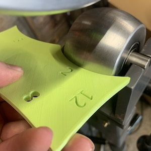 May include: A lime green plastic template with the numbers 12 and 2 printed on it. The template is being held up to a metal roller.