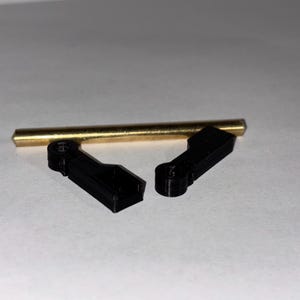 May include: An overhead shot shows a brass rod and two black plastic components. The rod is cylindrical and has a gold color. The black components are rectangular with a curved section. The items are arranged on a white surface.
