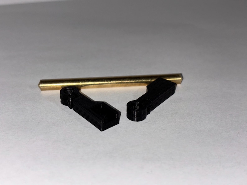 May include: An overhead shot shows a brass rod and two black plastic components. The rod is cylindrical and has a gold color. The black components are rectangular with a curved section. The items are arranged on a white surface.